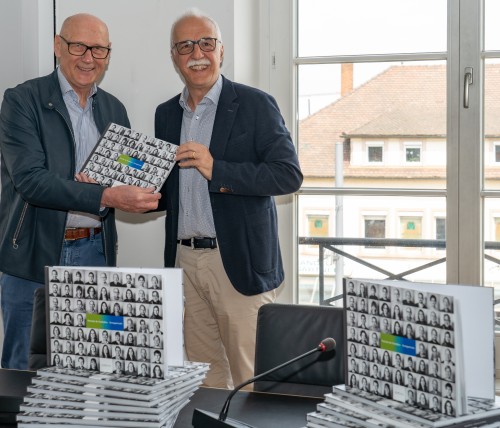 Hans-Werner Hilzinger, der Geünder der Rheinstiftung, und der ehemalige OB Toni Vetrano halten gemeinsam eine Ausgabe des Buches Grenzportraits in die Kamera. Vor ihnen liegt ein Stapel des Buches Grenzportraits. 