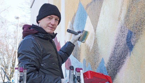 Künstler Johannes Mundinger steht mit Pinsel vor der Freibadwand.