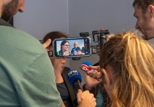 Calorie stellt sich vor zwischen den von hinten aufgenommenen Journalisten, sieht man das Bild der Generaldirektorin auf dem Handy, mit dem sie gefilmt wird.