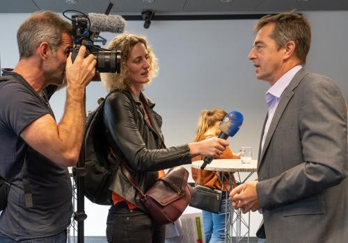 Kameramann und Journalistin mit Mikro, beide links stehend, mit dem sprechenden Harald Höflich gegenüber                                              