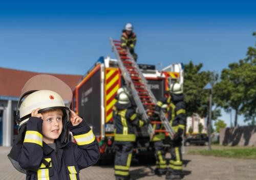 ein kleiner Junge in viel zu großer Feuerwehrkleidung mit Helm im Vordergrund; im Hintergrund lassen Feuerwehrleute eine Leiter von einem Feuerwehrfahrzeug herunter                                                               