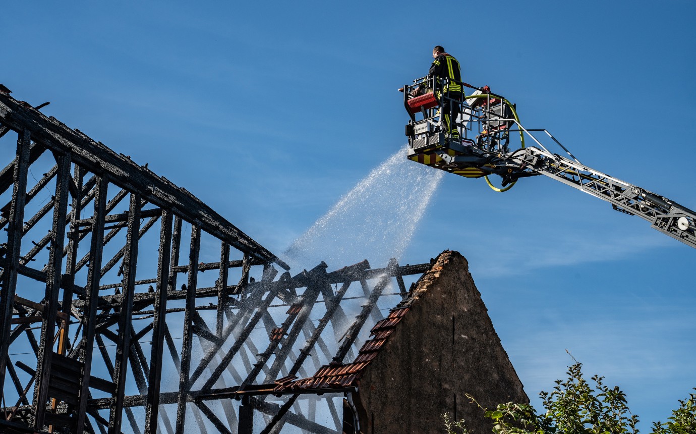 Ein Feuerwehrmann spritzt aus dem Korb der Drehleiter durch das verkohlte Dachgebälk große Mengen von Wasser in die Brandruine                             
