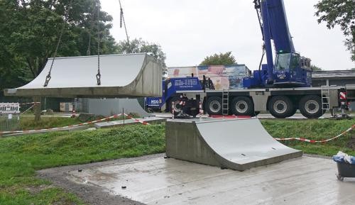 Ein Teilstück der Halfpipe hängt an Ketten über der Betonbodenplatte und wird von einem Autokran gehalten.