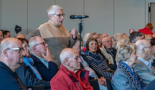Eine Frau steht hinter einem Mikrofon, umringt von sitzenden Zuhörerinnen und Zuhörern.
